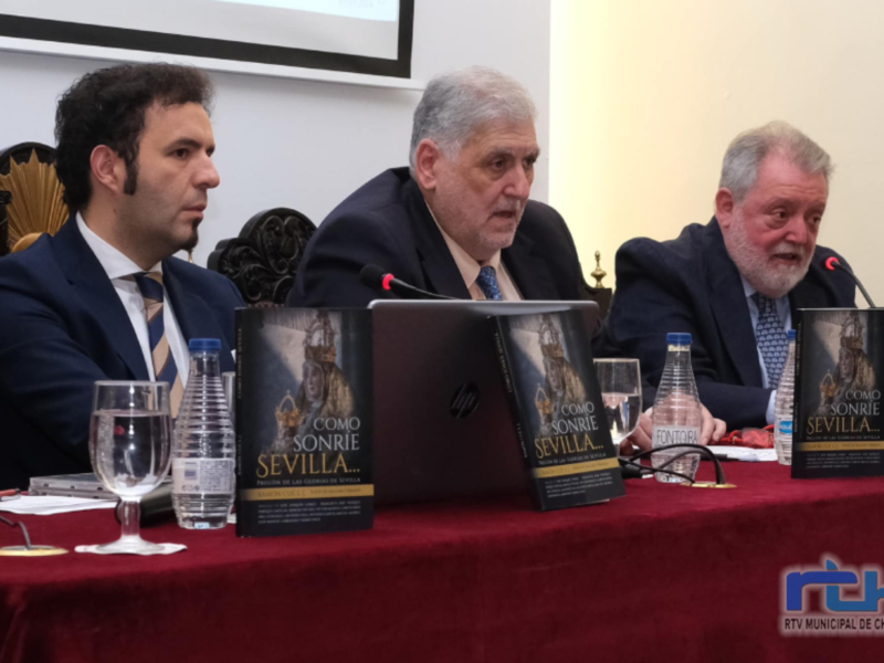 Tres hombres sentados en una mesa con libros titulados 'Cómo sonríe Sevilla' en una conferencia o presentación.