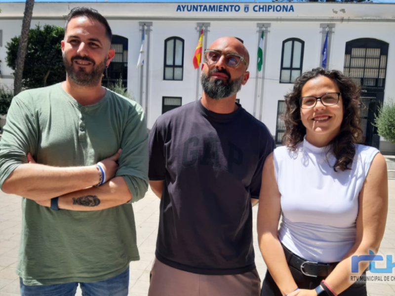 Tres personas sonrientes frente al Ayuntamiento de Chipiona, con banderas y árboles al fondo.