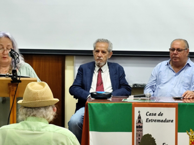 Una conferencia en la Casa de Extremadura, con tres personas en el escenario, una de ellas con un sombrero, y una bandera de Extremadura en el fondo.
