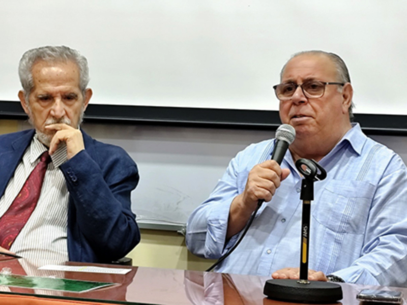 Dos hombres en una conferencia de prensa, uno con micrófono y el otro con una hoja de papel en la mano.