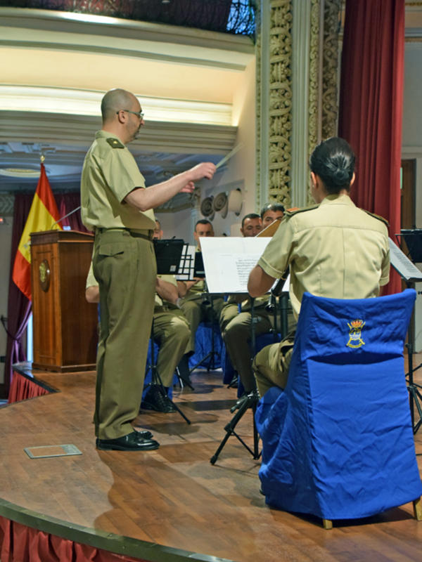 Imagen de un hombre en uniforme militar dando una presentación a otros militares sentados. El fondo es una sala de conferencias con un escenario, cortinas rojas y una bandera.