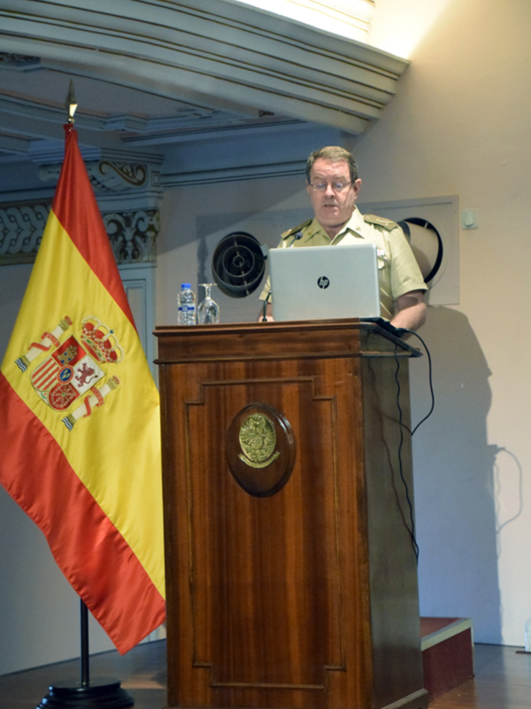 Un hombre en uniforme militar está en un podio con una bandera española a su lado, usando un portátil HP.
