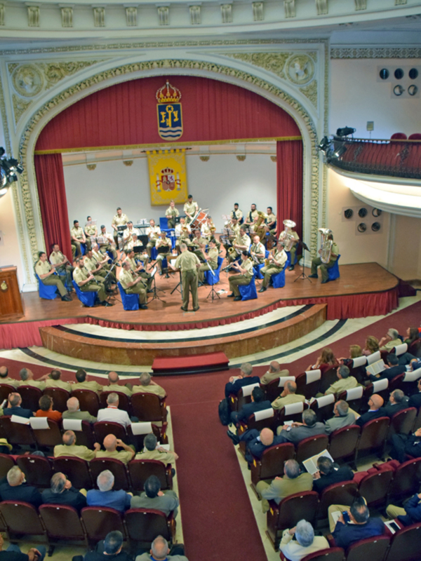 Una banda militar toca en un escenario de teatro con una coronación real en el fondo. La audiencia se sienta en filas de asientos rojos y marrones, observando la actuación. La decoración del teatro es elegante con detalles dorados y rojos, creando un ambiente formal.