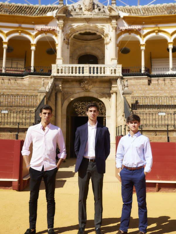 Tres jóvenes hombres posan frente a la famosa Plaza de Toros de la Real Maestranza de Sevilla, España, con el techo azul y la arquitectura histórica en el fondo.