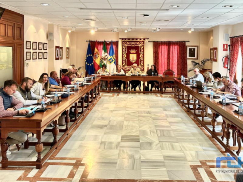 Sala de reuniones con personas sentadas en mesas, banderas y decoración oficial en el fondo.