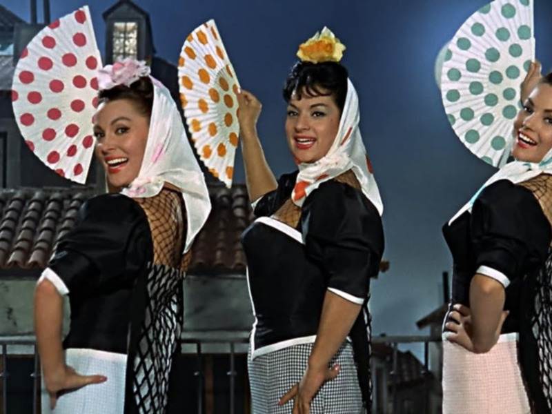 Tres mujeres en trajes de camareras con pañuelos y abanicos, posando con alegría en un escenario urbano.
