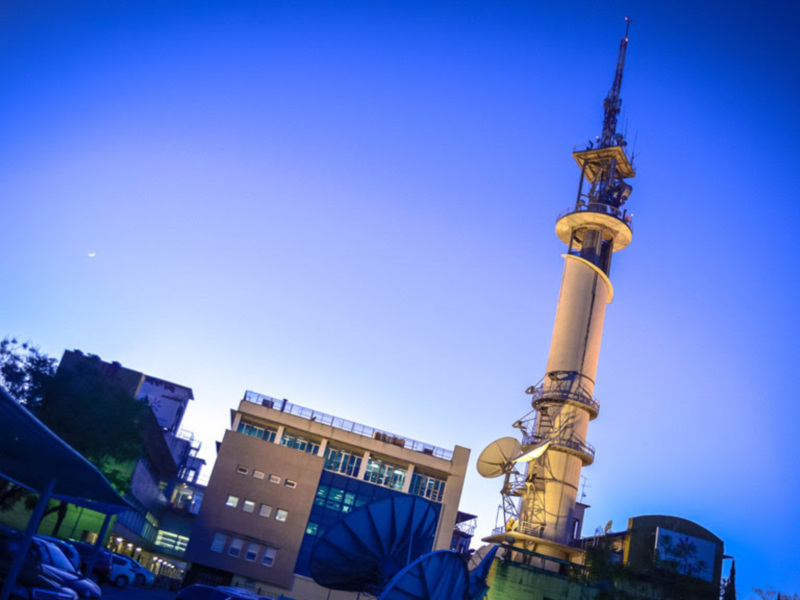 Torre de telecomunicaciones al atardecer con edificios urbanos en el fondo.