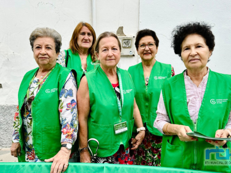 Grupo de voluntarias en chalecos verdes, posando frente a una fachada blanca, con una mesa verde en el frente.