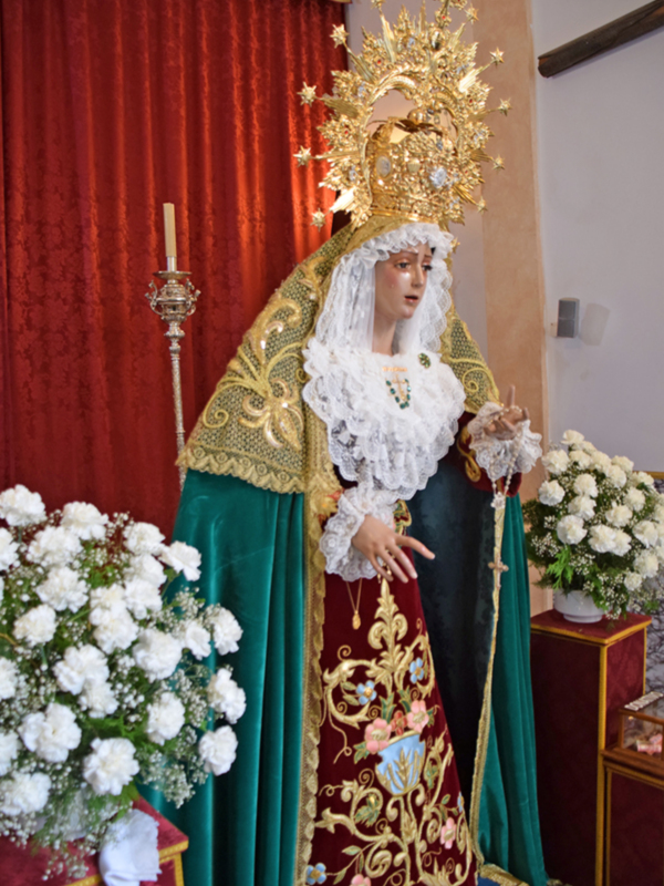 Estatua de la Virgen María con vestimentas ornamentadas y corona dorada, rodeada de flores blancas en un entorno religioso.