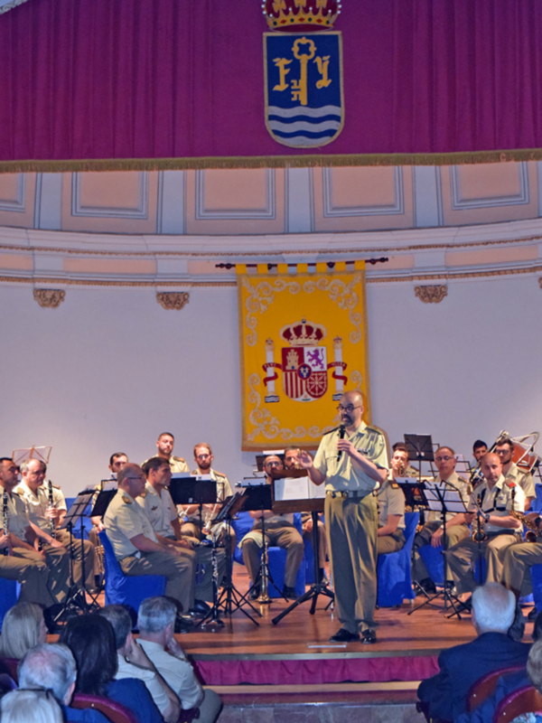 Concierto de banda militar en un escenario formal con escudo real en el fondo.