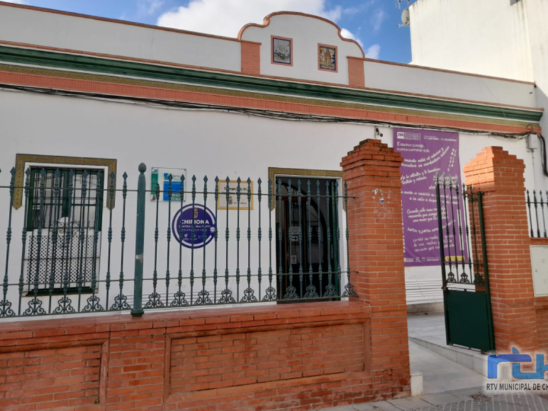 Fachada de un edificio con ventanas y puerta principal, junto a una cerca metálica y un cartel informativo en la pared.