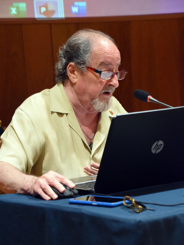 Un hombre con gafas y una camisa beige está usando un ordenador portátil HP en una conferencia. En el fondo, se puede ver una pantalla con iconos de Microsoft Office.
