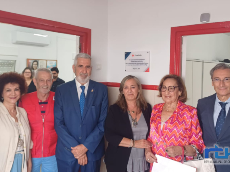 Entrega de un cheque en un evento de Cruz Roja, con personas en un ambiente formal. La imagen muestra una transacción financiera entre dos personas, con un fondo que incluye elementos de decoración y un cartel de Cruz Roja en el fondo.