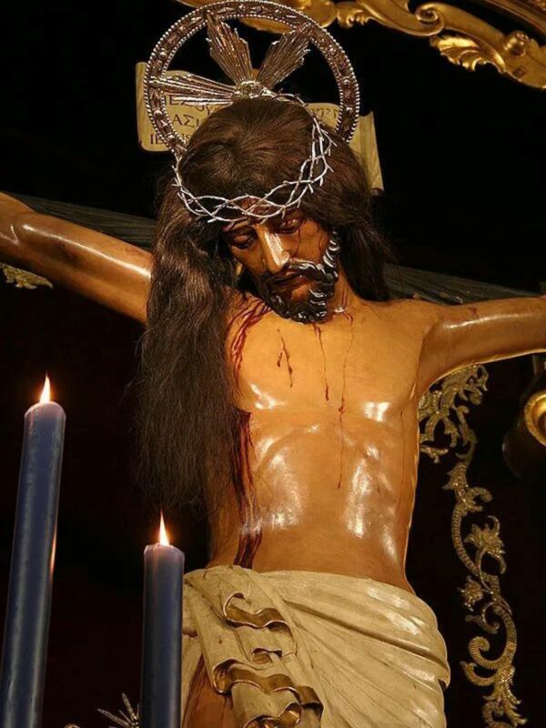 Estatua religiosa de Cristo crucificado con corona de espinas y sangre en la frente, rodeada por velas azules en una escena de penitencia religiosa.