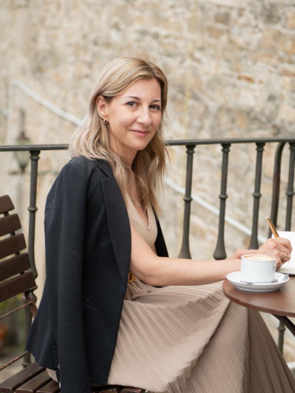 Una mujer disfruta de un café en un balcón con una vista de una antigua estructura de piedra.