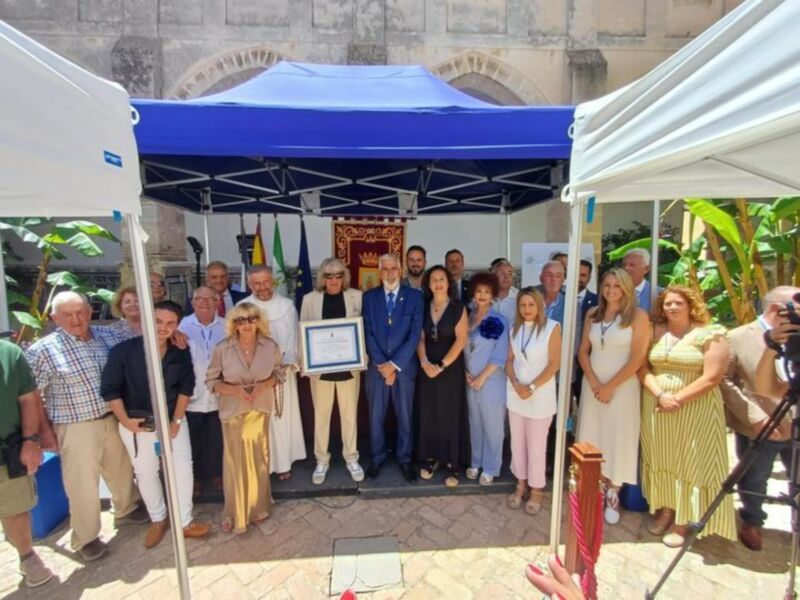 Celebración de un evento con una persona sosteniendo un certificado bajo una estructura de toldo en un entorno urbano con edificios históricos al fondo.