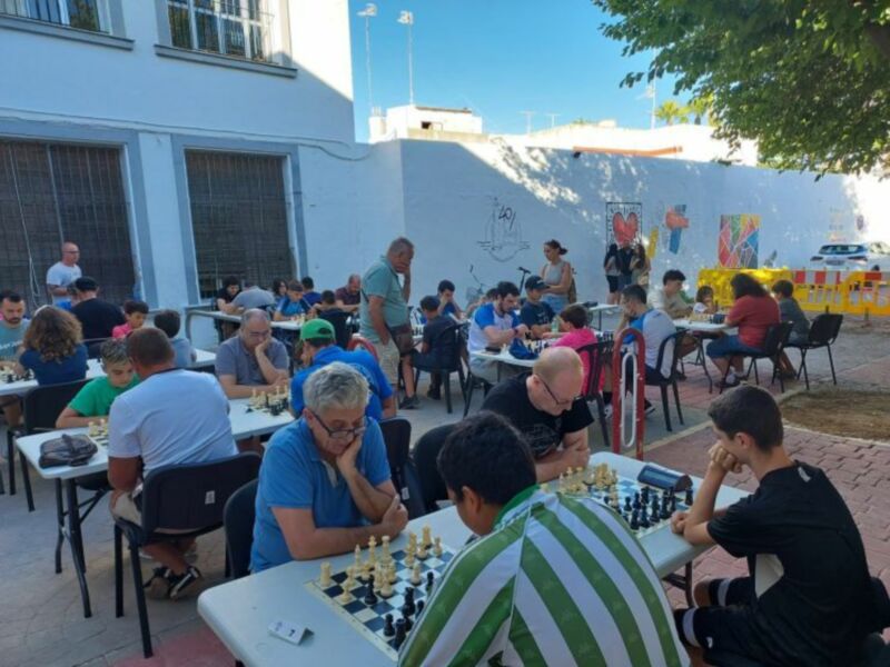 Jugadores de ajedrez disfrutan de un torneo al aire libre bajo el sol, rodeados de murales coloridos en un patio.
