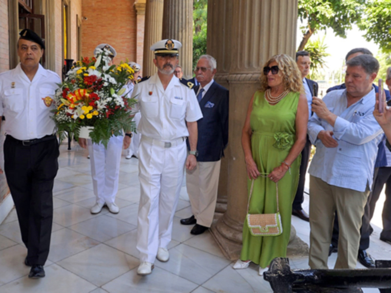 Un hombre en uniforme militar camina con un ramo de flores, seguido por una mujer en vestido verde y otros individuos en la calle. El ambiente parece ser formal o ceremonial.