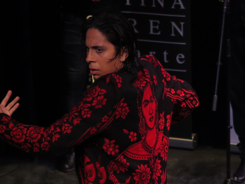 Una persona con una bufanda roja y negra con un diseño floral, posando en un evento con un fondo negro y letras blancas.
