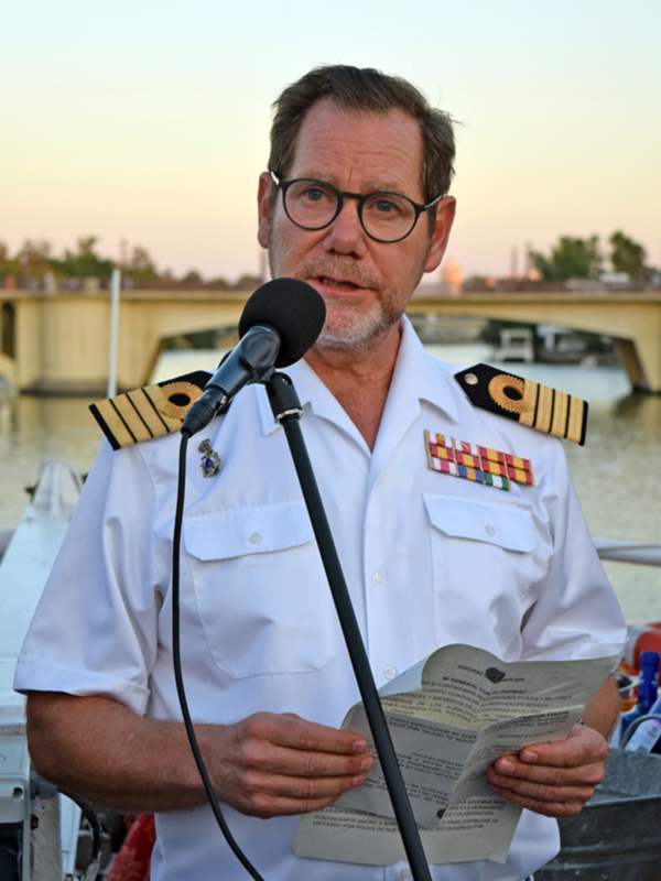 Oficial naval hablando en un evento al aire libre frente a un puente y agua.