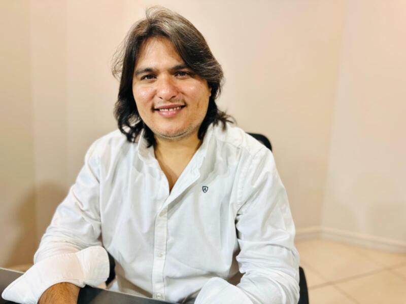 A man with long hair and a white shirt, sitting in an office setting.