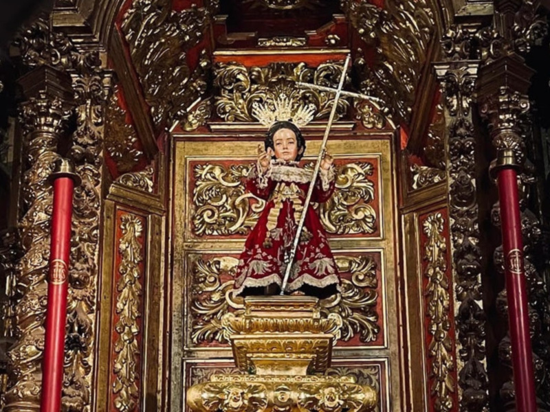 Elegante estatua religiosa en un altar decorado con detalles dorados y rojos, iluminada por una vela roja.