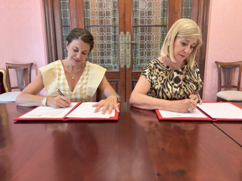 Damas firmando documentos en una mesa con un fondo de puertas de madera y vidrio decorativo.