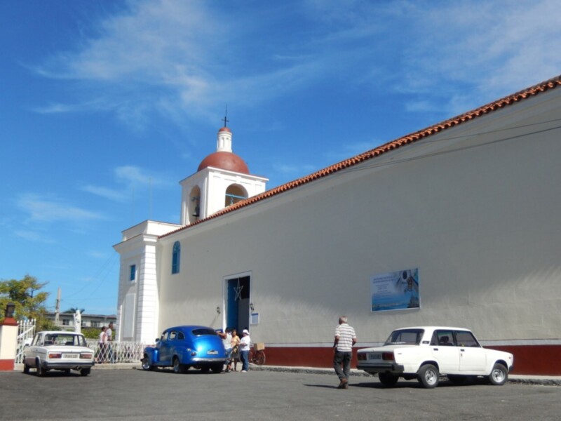 Iglesia histórica con cúpula roja y fachada blanca, rodeada de coches clásicos y personas en un día soleado.