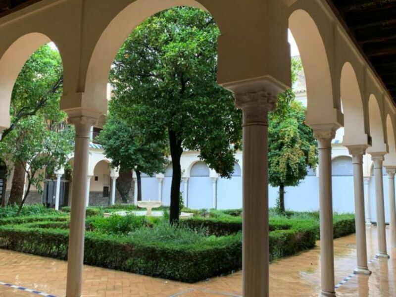 Jardín interior con arcos y columnas blancas, rodeado de arbustos y árboles verdes.