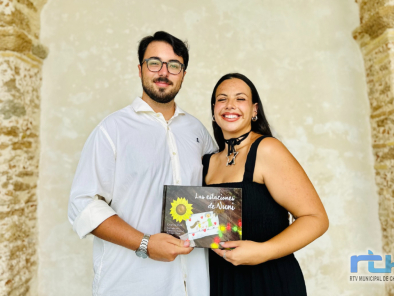 Un hombre y una mujer sonrientes, posando con un libro titulado 'Las estaciones de Nuni' en un entorno histórico con una torre de ladrillo en el fondo.