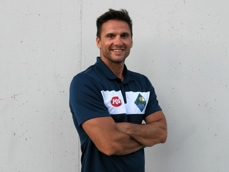 Persona sonriente en camiseta PQS, con brazos cruzados, frente a pared blanca.