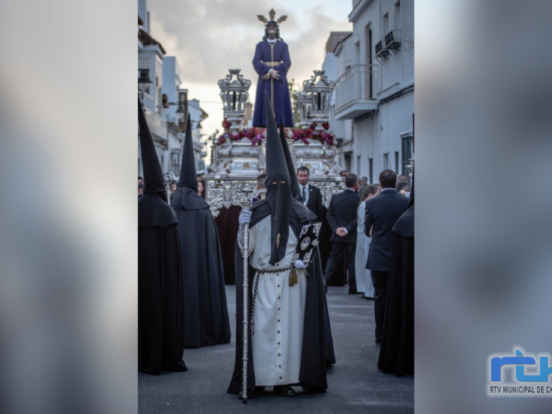 Procesión religiosa con personajes en trajes tradicionales y una estatua en el centro, en un entorno urbano con edificios blancos y azules.