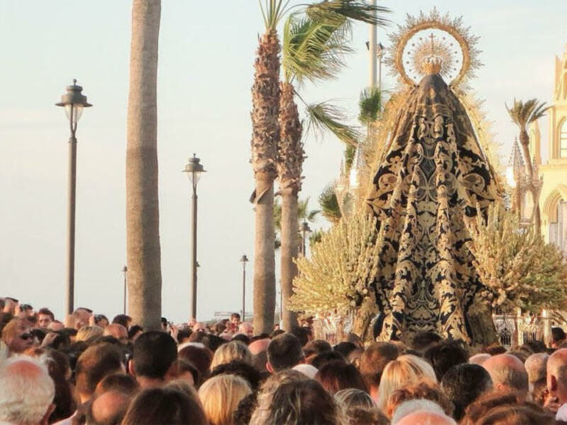 Procesión religiosa con una imagen venerada en el centro, rodeada por una multitud de fieles, con palmeras y edificios históricos al fondo.