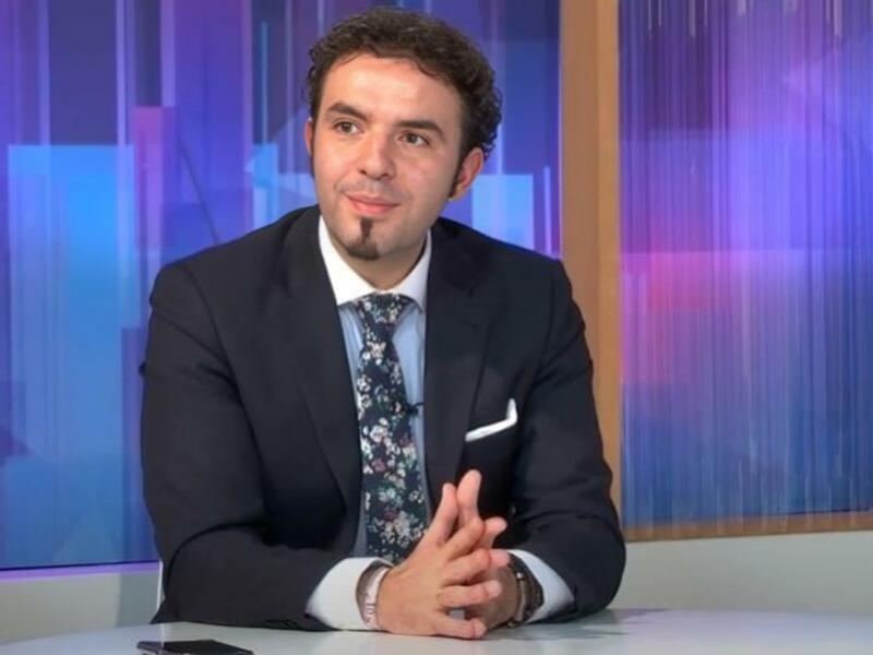 Un hombre en traje y corbata floral, sentado en un estudio de televisión con un fondo de colores azules y púrpuras.