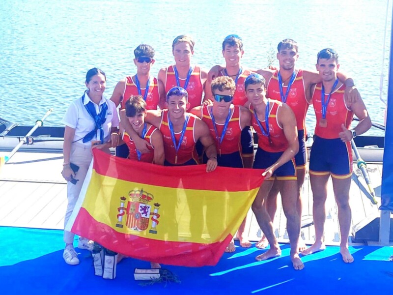 Equipo español de remo con medallas y bandera española en un evento deportivo.