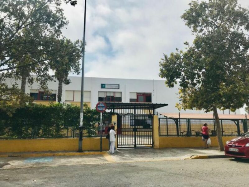 Entrada principal de un edificio blanco con ventanas y puertas rojas, rodeado de árboles y una valla amarilla.