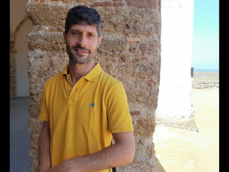 Hombre en polo amarillo frente a una pared de ladrillo en un entorno al aire libre.