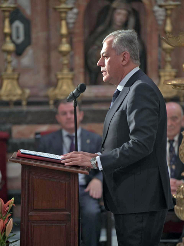 Hombre en traje hablando en un podio en un entorno formal, posiblemente una ceremonia o evento oficial.