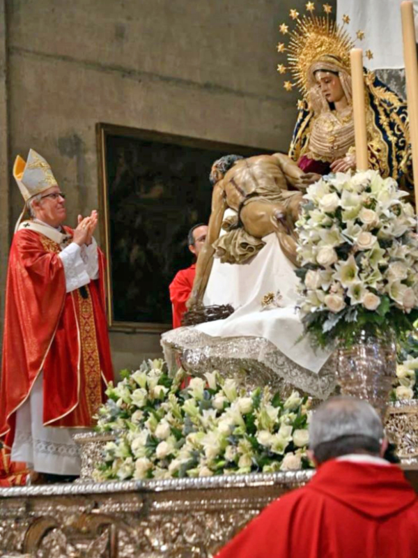 Especial momento religioso en una iglesia, con una figura vestida con un manto y tocado con un halo dorado, rodeada de flores blancas y un obispo en el fondo, celebrando una ceremonia importante.