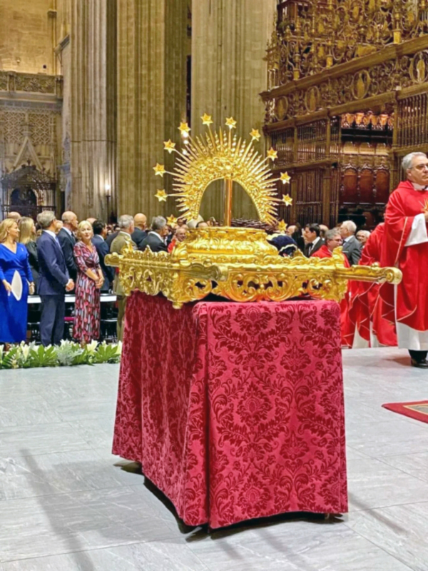 Imagen de una procesión religiosa en una iglesia, con un altar dorado y una tela roja en el fondo.
