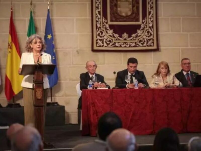 Una conferencia de prensa en un salón con una mujer en el centro del escenario, flanqueada por tres hombres sentados en una mesa con banderas en el fondo.