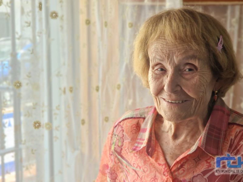 Retrato de una anciana sonriente, vestida con una camisa de estampado floral, frente a un fondo de cortinas transparentes con patrones florales.