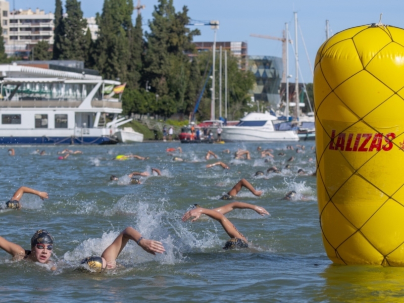 Competidores nadando en una competencia de natación en aguas abiertas con un marcador flotante amarillo grande en el centro.