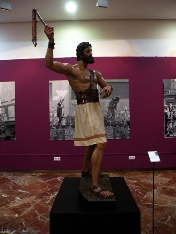 Una estatua de un hombre con una correa en la mano, exhibida en un museo. La estatua está sobre una plataforma negra y está iluminada por luces de techo. En el fondo, hay dos fotos de personas con sombreros y una pared de color púrpura.