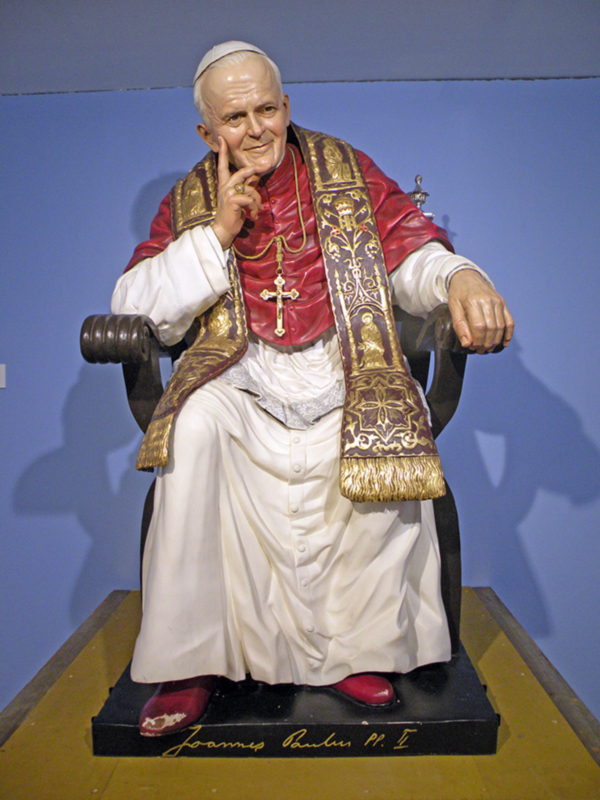Estatua del Papa Juan Pablo II, vestido con su traje sacerdotal rojo y blanco, sentado en un trono.