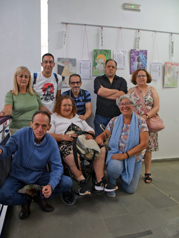 Grupo de personas en una exposición de arte, con carteles colgados en la pared. El ambiente es informal y amistoso, con personas de diferentes edades posando para la foto.