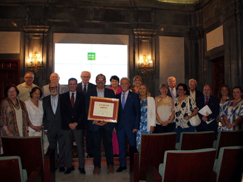 Grupo de personas posando para una foto en un salón con sillas y un diploma en mano.