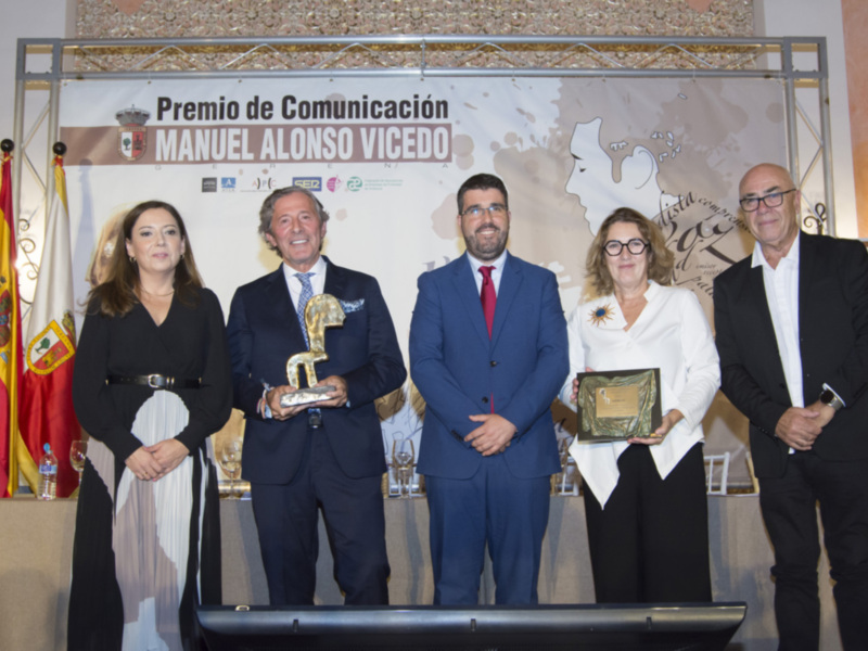 Premio de Comunicación Manuel Alonso Alonso Vicedo en un evento formal con personas en traje y una bandera española en el fondo.
