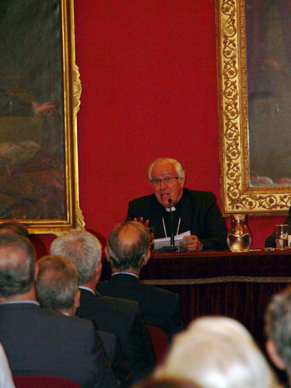 Un clérigo habla en un evento formal en un salón con decoración clásica y asistentes atentos.
