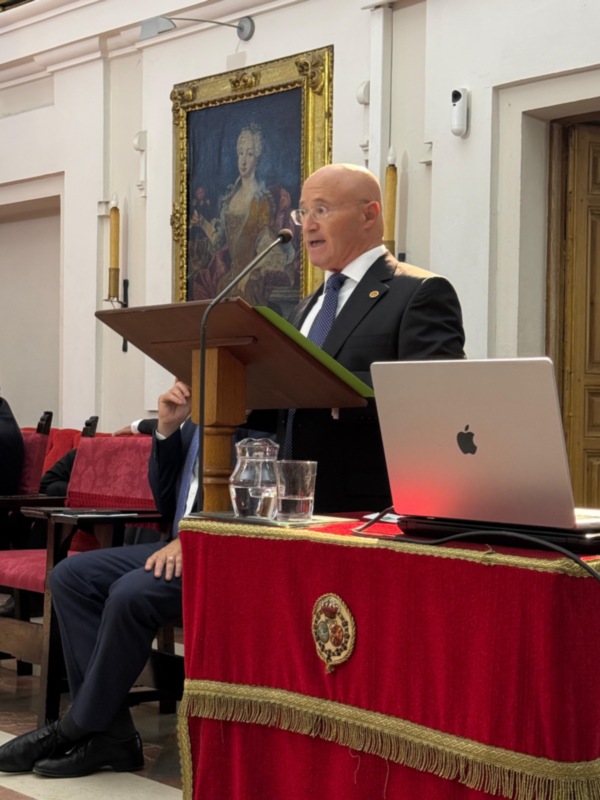 Hombre hablando en un podio en un salón con pintura histórica en el fondo.